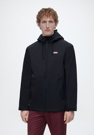 Mees lokkis punaste juustega, kandes musta kapuutsiga jope, mis on lukuga suletud, ning marooni värvi pükse, seisab lihtsa heleda tausta ees.