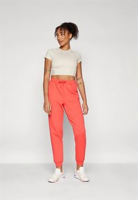 Moteris su garbanotais plaukais, apsirengusi baltu trumpu marškinėliu, koralinėmis sportinėmis kelnytėmis ir baltomis sportinėmis bateliais, stovi prieš paprastą šviesų foną.