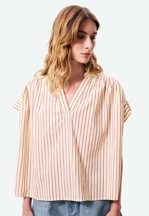 Femme blonde portant une blouse ample crème à rayures rouges verticales et col en V, associée à un pantalon bleu texturé, regardant vers le bas sur le côté.