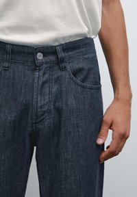 Ingrandimento della vita di una persona che indossa pantaloni di jeans scuri con una semplice maglietta bianca, mostrando la texture del tessuto e la chiusura con bottone.