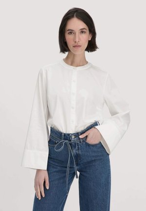 Witte longsleeve shirt met een ronde halslijn en knoopsluiting, gecombineerd met high-waisted blauwe jeans met een gestrikt taille detail.