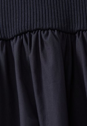 Femme aux cheveux roux portant une robe maxi noire sans manches et des chaussures beiges, debout devant un fond uni gris clair.