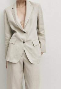 Blazer en lin beige clair avec un décolleté en V profond, deux poches frontales et des boutons noirs, assorti à un pantalon ample assorti.