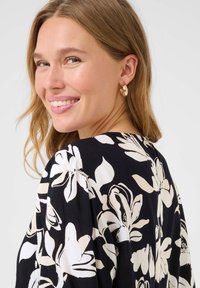 Blouse en tissu noir avec un motif floral blanc, col rond, texture douce et manches courtes, accompagnée de boucles d'oreilles en or. 