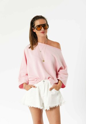 Femme portant un pull oversize rose épaules dénudées, un short blanc effiloché, de grandes lunettes de soleil teintées et des colliers en or superposés, les mains dans les poches.