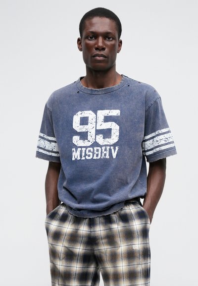 Plavi, veliki pamučni T-shirt s iznošenim rubovima, na kojem se nalazi veliki bijeli natpis "95" i "MISDHRV". Uparen s ležernim hlačama u kariranom uzorku.