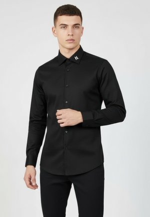 Ung mand iført en tætsiddende sort skjorte med knapper og et lille hvidt logo på kraven, stående foran en ensartet lys baggrund.