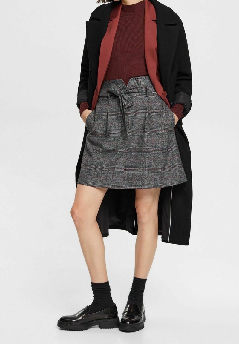 Femme portant un manteau noir, un haut couleur rouille, une jupe grise taille haute avec ceinture, des chaussettes noires et des mocassins noirs brillants, debout les mains dans les poches.