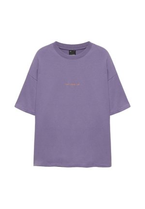 Jeune homme portant un t-shirt violet oversize, un long short en jean bleu clair, des chaussettes blanches et des baskets vert olive, debout contre un mur blanc.