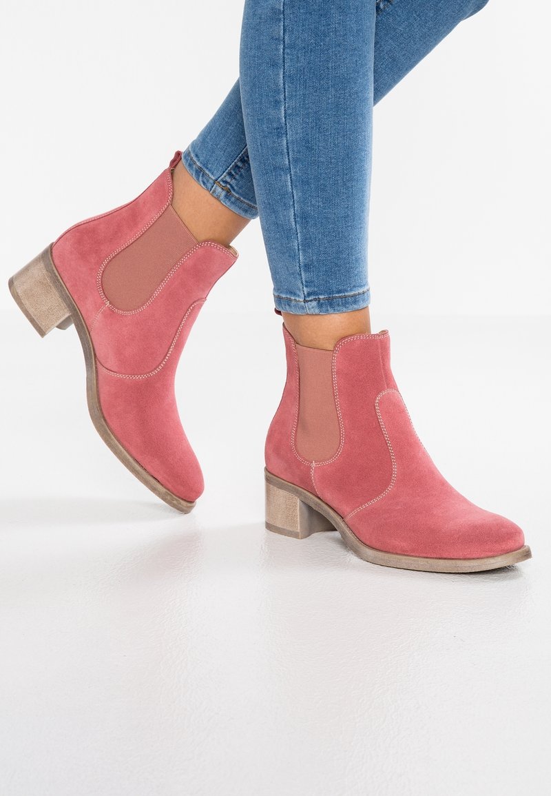 Personne portant des bottines roses en daim avec des talons blocs bas et un jean bleu sur fond blanc.