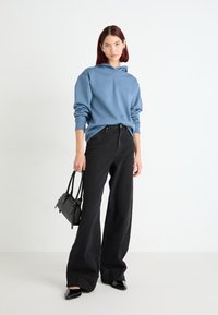 Světle modrá mikina s kapucí, černé kalhoty s širokým střihem, černá kabelka a špičaté boty. Hladká textura, volný střih a minimalistický design.