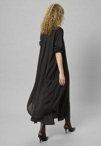 Femeie cu părul creț care se îndepărtează, purtând o rochie lungă neagră, colanți negri transparenți și pantofi cu toc înalt negri, pe un fundal gri uniform.