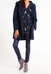 Parka bleu marine avec capuche, haut à motifs gris et blancs, pantalon bleu marine, et bottines grises claires avec fermetures éclair. Textures lisses partout.