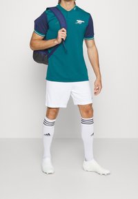 Polo bleu sarcelle avec manches bleu marine, short blanc, chaussettes rayées noires et blanches, et chaussures à crampons blanches. Porte un sac à dos bleu foncé.