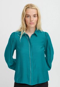 Camicia blu verde, con bottoni e maniche a sbuffo, dalla vestibilità dritta. Presenta un bordo decorativo lungo la linea dei bottoni. Tessuto morbido e liscio.