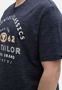 T-shirt bleu marine à manches courtes en tissu texturé, avec du texte blanc et or. Le design comprend un logo circulaire et des détails de marque.