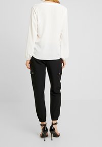 Donna che indossa una camicia bianca a maniche lunghe, pantaloni cargo neri con tasche laterali e sandali con tacco alto in vernice nera, in piedi.