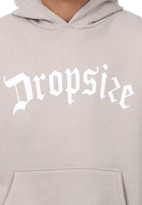 Kapuzensweatshirt aus hellgrauem Stoff mit einer Fronttasche und großen weißen Buchstaben, die "Dropsize" in einer stilisierten Schriftart lesen.