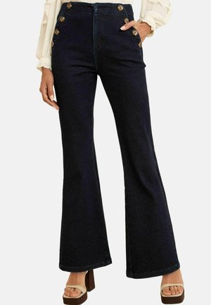 Jeans de cintura alta acampanados en denim oscuro con detalle de botones laterales, que presentan una textura suave y costuras prominentes en los bordes.