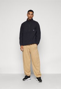 Zwart fleece pullover met een kwart rits, voorzien van een contrasterend paneel; gecombineerd met lichtbeige, taps toelopende broek en zwarte sneakers.