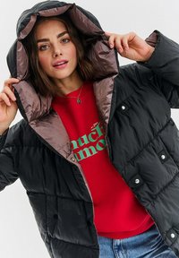 Puchowa czarna kurtka z błyszczącą brązową podszewką, z zatrzaskami i przypinaną kapturem, noszona na czerwonym swetrze z zielonym napisem.