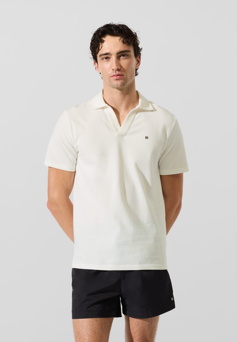 Camiseta polo crema de manga corta y textura suave. Presenta una tapeta de dos botones y un pequeño logo negro en el lado izquierdo del pecho.