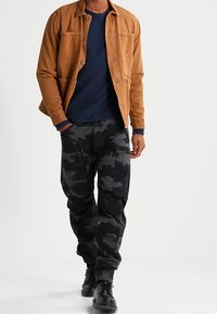 Uomo che indossa una giacca marrone, un maglione blu navy, pantaloni cargo mimetici neri e grigi e stivali neri con lacci mentre cammina in avanti.