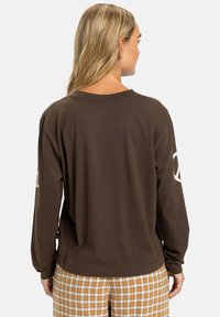 Camicia a maniche lunghe marrone con vestibilità rilassata, caratterizzata da un colletto rotondo e grafiche di simboli della pace di colore chiaro sulle maniche.
