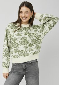 Pull en maille avec un motif floral vert sur fond crème, poignets et ourlet côtelés, coupe décontractée, associé à un jean en denim gris.