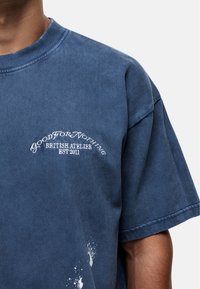 T-shirt di cotone blu con maniche corte, colletto rotondo e stampa sul petto anteriore in testo bianco. Dettagli sbiaditi simili alla vernice lungo l'orlo.