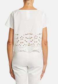 Hvid kortærmet top med en gennemsigtig ryg, der har intrikate broderier og udskårne blomsterdesign, bølget kant og en afslappet pasform.