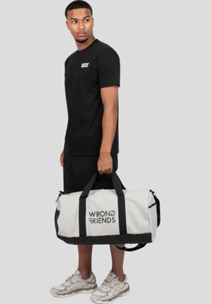 White duffel bag with black straps and "WRONG FRIENDS" logo; person wearing black shirt and shorts, gray sneakers, standing against a gray background.