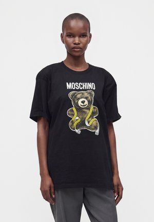 Žena s krátkymi vlasmi nosí čierne tričko Moschino so vzorom medvedíka zabaleného do žltej meter.