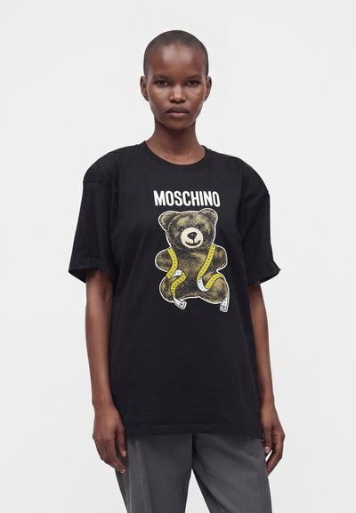 Γυναίκα με κοντά μαλλιά φορώντας μαύρο t-shirt Moschino με αρκουδάκι τυλιγμένο σε κίτρινη μεζούρα.