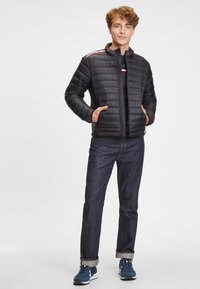 Černá puffová bunda se zipem vpředu, ribovanými rukávy a dvěma bočními kapsami. Obsahuje červené a bílé pruhy na rameni.