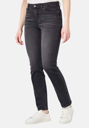 Frau trägt eine dunkelgraue, eng geschnittene Jeans und weiße Sneaker, steht vor einem einfarbigen Hintergrund, Hände entspannt an den Seiten.