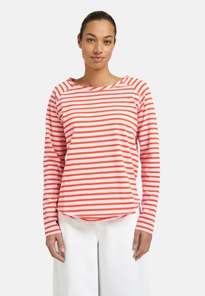 Woman with tied-back hair wearing a long-sleeve white and orange striped shirt and white pants, standing against a plain background.