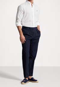 Biała lniana koszula zapinana na guziki, granatowe spodnie w prążki oraz granatowe espadryle z haftowanymi zdobieniami. Widoczne kieszenie, dopasowany krój.