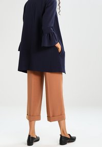 Osoba v námořnickém kabátu s rozevlátými rukávy, karamelových zkrácených kalhotách s širokými nohavicemi a černých nazouvacích botách, stojí s rukama v kapsách.