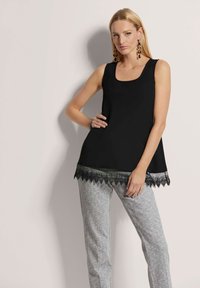 Schwarzes Tanktop mit quadratischem Ausschnitt und mit gewelltem Spitzenbesatz am Saum, kombiniert mit grauen gemusterten Hosen. Glatte Stofftextur.