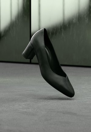 Chaussure à talon moyen en cuir noir flottant au-dessus d'un sol moquetté gris avec un arrière-plan vert flou.