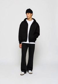 Jonge man met zwarte beanie, zwarte zip-up hoodie, wit T-shirt, zwarte broek en witte sneakers, staand tegen een effen witte achtergrond.