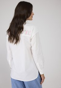 Weißes Baumwollhemd mit mehrfarbiger "LOVE"-Stickerei auf dem Rücken, langen Ärmeln und abgerundetem Saum. Lockerer Schnitt mit Knopfmanschetten.