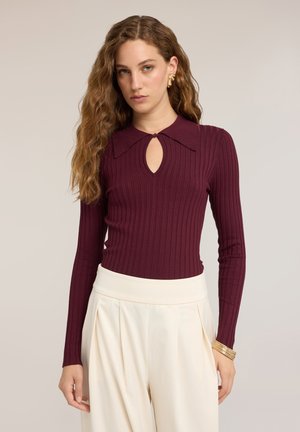 Donna con capelli lunghi e mossi indossa una maglia a coste bordeaux a maniche lunghe con scollo a goccia e pantaloni bianchi a vita alta e pieghettati.