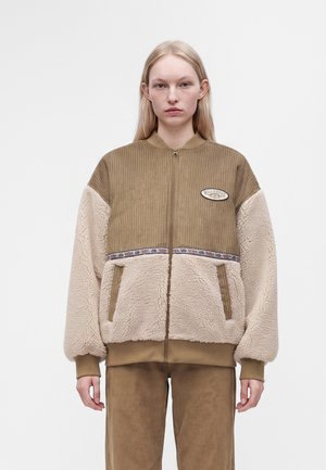 Femme portant une veste polaire marron et beige avec fermeture éclair à l'avant et un pantalon marron assorti, debout devant un fond blanc uni.