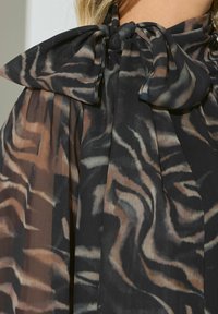 Schwarz-beige gemusterte Bluse mit großem Schleifenkragen, die aus einem transparenten, strukturierten Stoff gefertigt ist und einen fließenden Schnitt hat.