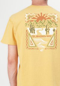 Gul t-skjorte med grafisk design av fjell, kaktuser og sol. Designet inkluderer jordfarger og geometriske mønstre.