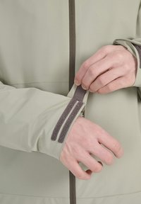 Hands adjusting the Velcro cuff strap on a light green jacket sleeve with a front zipper closure.