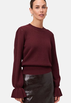 Femme portant un pull textured à manches longues couleur bordeaux avec des poignets plissés et une jupe en cuir ajustée sur un fond uni.