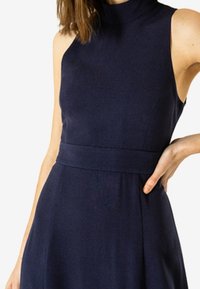 Námořnická modrá bezrukávová šaty s vysokým výstřihem, přesně padnoucí korzet a detail pasu. Vyrobené z hladké, pružné tkaniny s jemným leskem.